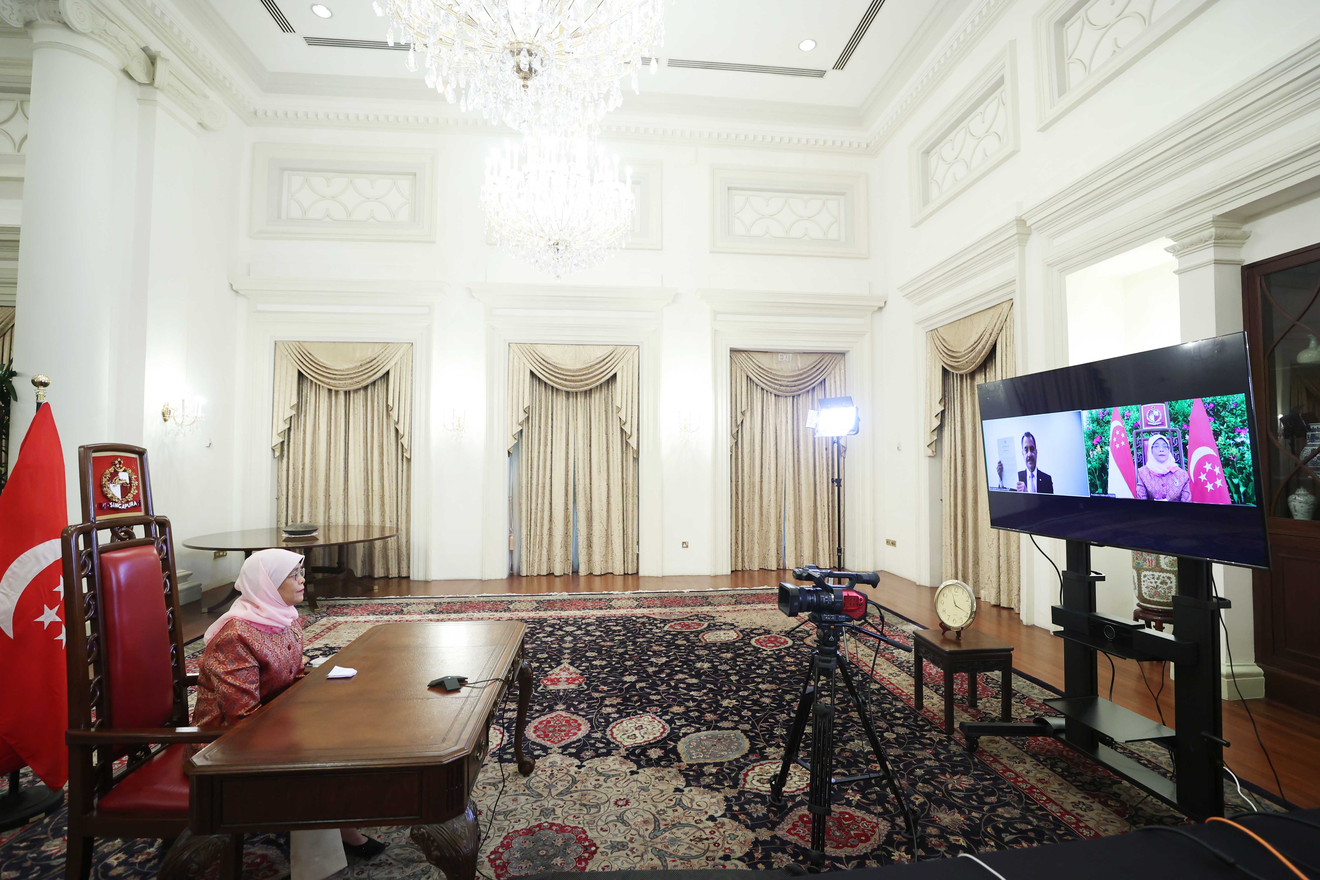 Halimah Yacob in ornate room, video conferencing on a large TV with Singapore flag.